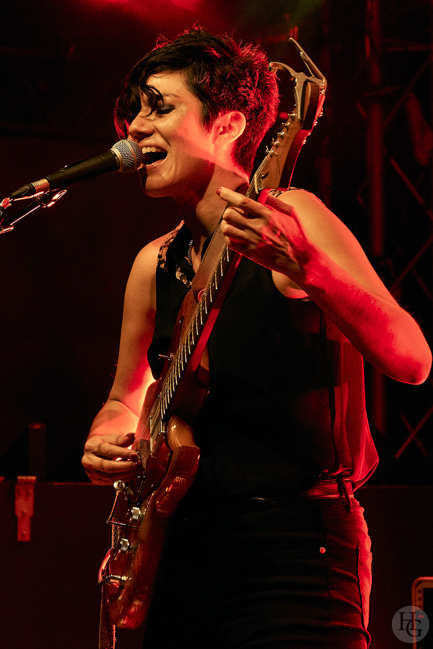 Ava Mendoza performs on a stage lit by red lights. Visible from the hips up, they are turned to the left and play an electric guitar while singing into a microphone, eyes closed. They are wearing black jeans with a black belt, a black sleeveless button up top with leopard print highlights on the shoulders, and a silver chain necklace.