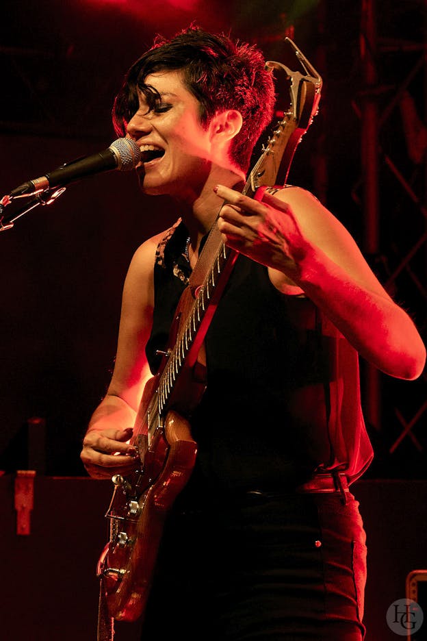 Ava Mendoza performs on a stage lit by red lights. Visible from the hips up, they are turned to the left and play an electric guitar while singing into a microphone, eyes closed. They are wearing black jeans with a black belt, a black sleeveless button up top with leopard print highlights on the shoulders, and a silver chain necklace.