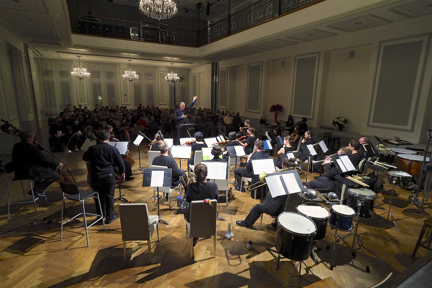A photograph of Petr Kotik performing, conducting a large ensemble in front of him. Behind him, a crowd of audience members watch. The room is high ceilinged with four chandeliers, white walls, and light blue panels. 