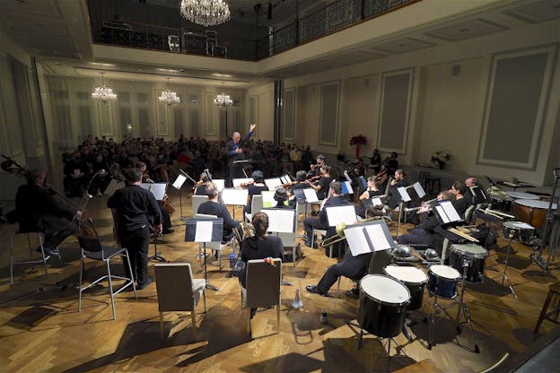 A photograph of Petr Kotik performing, conducting a large ensemble in front of him. Behind him, a crowd of audience members watch. The room is high ceilinged with four chandeliers, white walls, and light blue panels. 