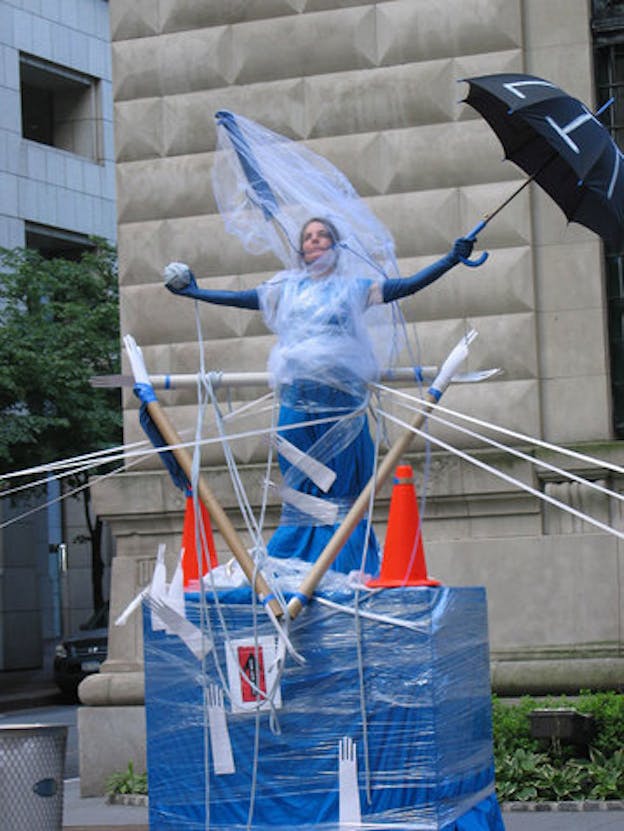 Mac Low performs in a city street. She is wrapped in white tulle and blue plastic and stands on top of a box wrapped in the same material. In one hand she holds a ball of yarn and in the other hand, she holds a black umbrella with 