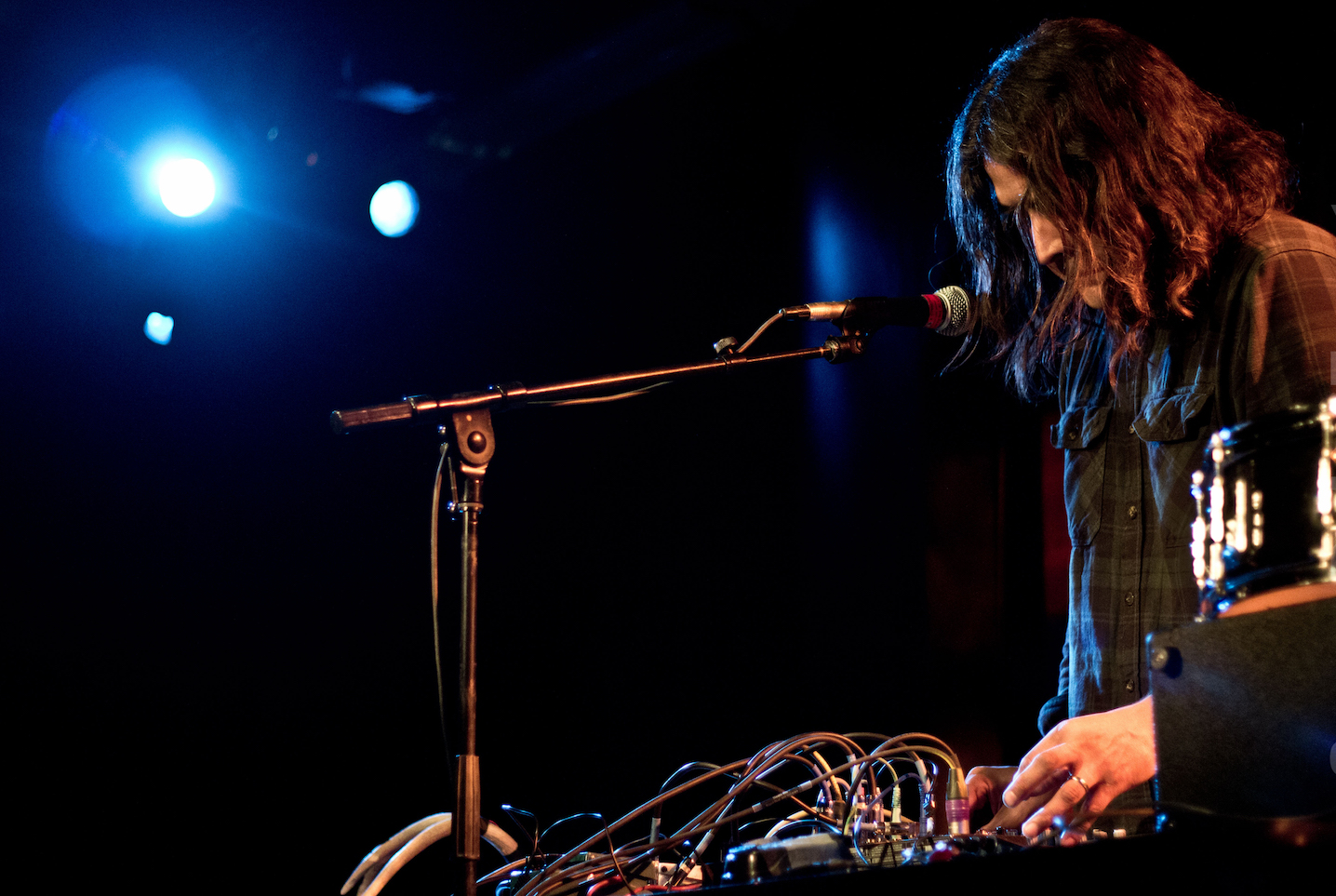 Raven Chacon performs, leaning over a sound box with many multi colored wires attached to it. A microphone is angled towards his mouth and he is illuminated by bright blue light in an otherwise dark setting. 