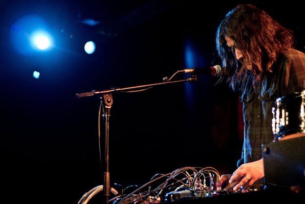 Raven Chacon performs, leaning over a sound box with many multi colored wires attached to it. A microphone is angled towards his mouth and he is illuminated by bright blue light in an otherwise dark setting. 