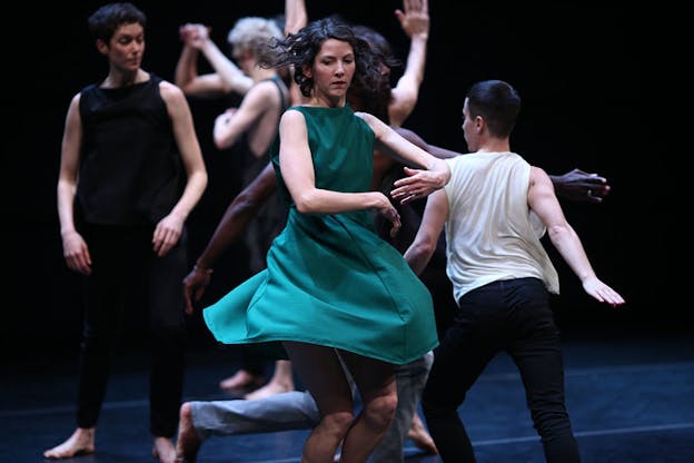 A performer wearing a green dress spins in the center. Behind them, there are several other performers wearing black and white. Some dance and hold hands. Others lunge and extend their arms beside them. One performer stands upright and looks at the others.
