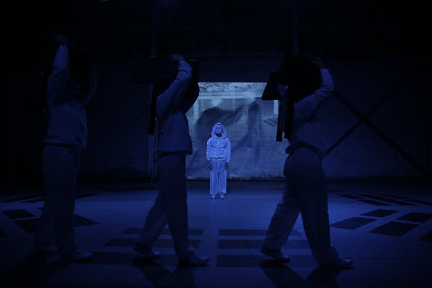 Three performers process across a darkened stage from left to right, silhouetted and holding their hands above their heads. Behind them, a performer dressed in white pants, white hooded sweatshirt, and white facemask faces the camera, well-lit and in striking contrast to the other three. A video of two blurry figures is projected against the back wall and the whole stage is bathed in blue light.