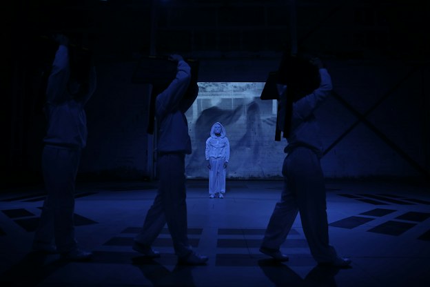 Three performers process across a darkened stage from left to right, silhouetted and holding their hands above their heads. Behind them, a performer dressed in white pants, white hooded sweatshirt, and white facemask faces the camera, well-lit and in striking contrast to the other three. A video of two blurry figures is projected against the back wall and the whole stage is bathed in blue light.