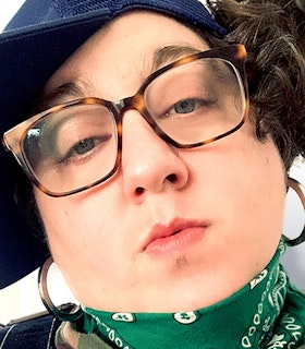 A close-up portrait of Constantina Zavitsanos. The artist wears tortuga glasses, hoop earrings, and a green bandana that covers their neck. 