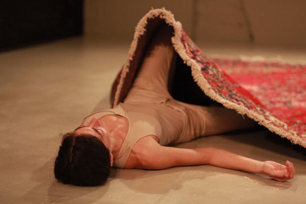 A performance still of Maria Hassabi wearing cream pants and a cream colored tank top. She lays on the floor, bending her left leg at the knee. A red carpet rests atop her legs. 