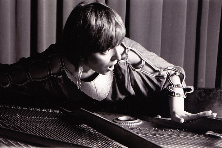 Grayscale photograph of a person with short light hair and a shirt with open loops on the sleeves laying on top of a sound machine.  
