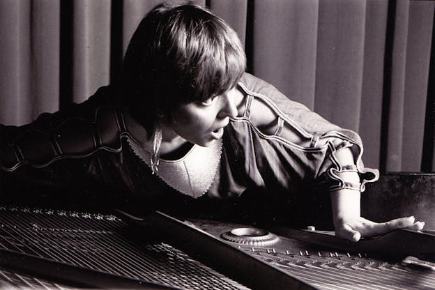 Grayscale photograph of a person with short light hair and a shirt with open loops on the sleeves laying on top of a sound machine.  