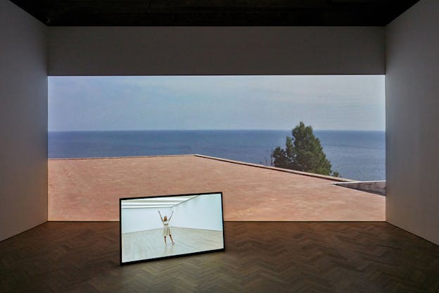On a wall the projection of a red terrace with the sea stands behind a small screen showing a figure in a bare room with their hands upwards.