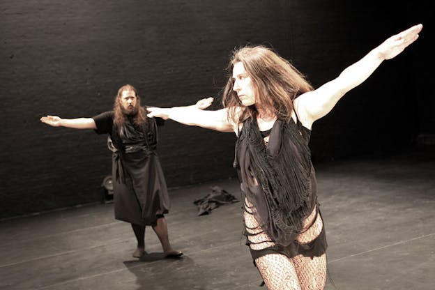 Performer dressed in sheer black fabric and lace tights looks sideways and extends both arms outwards while standing behind them in the black-walled and bright room, a person also dressed in black stares at them and extends both arms outwards.