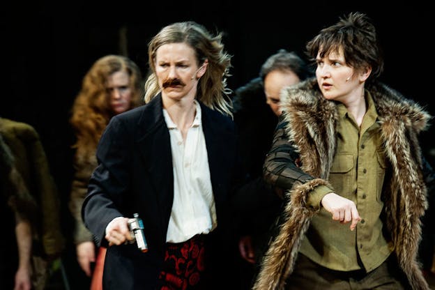 Close-up still of two performers in winter coats, one wearing a fake moustache and holding a handgun, walking together and looking intensely at something out-of-frame. Blurred behind them, three hunched performers follow.