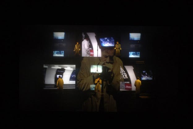 A performer wearing a yellow shirt talks closely into a microphone. Small projections of scenes are layered ontop of them in an otherwise black background.