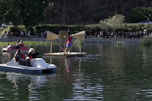 People seated on pedal boats surround in a lake a figure standing in between three big floating loudspeakers.
