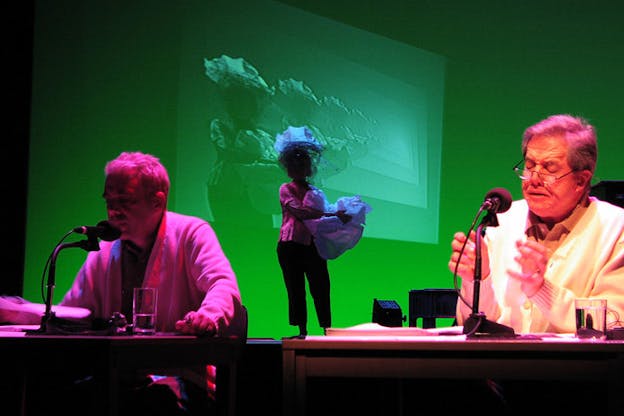 Two performers sit at desks and speak into microphones in the foreground. They are bathed in warm pink light. Behind them, a person holds fabric in front of a green-hued projection of their image which is copied and repeated many times.