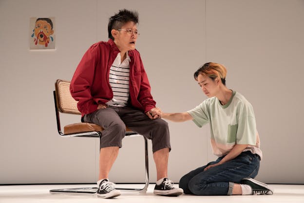 Aya Ogawa sits in a chair wearing glasses, a red jacket, a white and black striped polo shirt, capri pants, and low top sneakers. Ashil Lee kneels by their side, holding their hand in a light green and pink color blocked shirt with black pants and low top sneakers. A drawing of a crying child with a nosebleed hangs on the wall behind them.