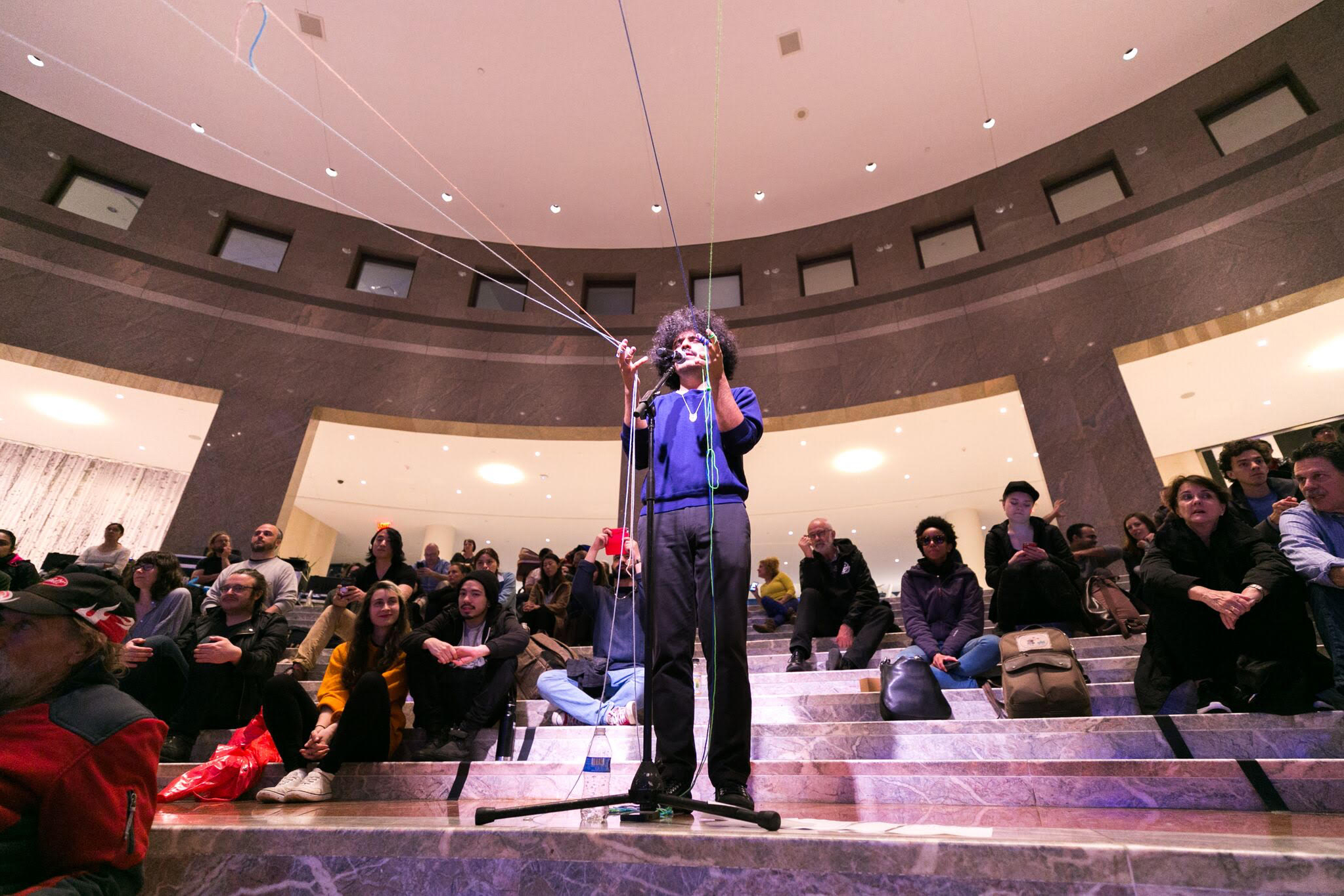 People seated on various levels of stairs surround a performer standing amidst them, singing in front of a mic and holding the hands up. Their fingers are interlaced in various colorful threads that are pulled in front of them.