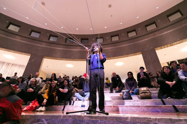 People seated on various levels of stairs surround a performer standing amidst them, singing in front of a mic and holding the hands up. Their fingers are interlaced in various colorful threads that are pulled in front of them.