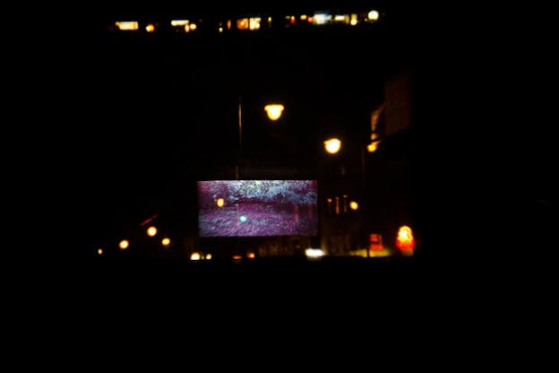 A small hanging screen projecting a blurred image of trees and dirt hued purple hangs in black space spotted with blurred, yellow and red lights evoking a city street at night. 