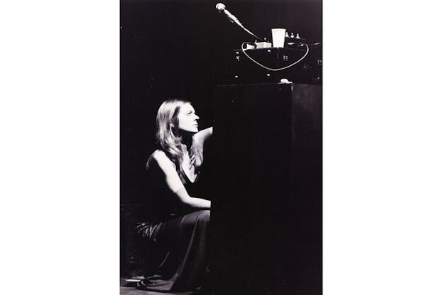 Black and white photograph of person with straight shoulder-length hair crouching down in front of a sound stand.