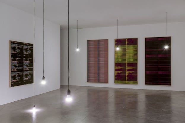 A gallery room with naked bulbs hanging from the ceiling almost down to the floor through black wires. On the two walls behind them rectangular canvases. On the left wall a canvas sits by itself in black, pink and blue colors while on the right wall three canvases sit aligned each with different width lines. The first canvas to the left in colors of pink and blue, the one in the middle in colors of green, yellow and magenta and the third hues of red magenta.