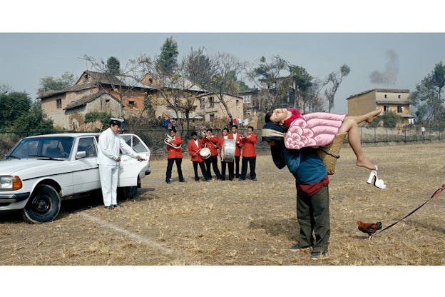 On the right, one performer carries Hawkins on their back in a basket. A rooster stands behind them. On the left, another performer dressed in white suit opens the rear door of a white car. In the middle back, a group of people dressed in red and black suits each plays a wind instrument or a percussion set. They are situated in a rural area with residences in the background.