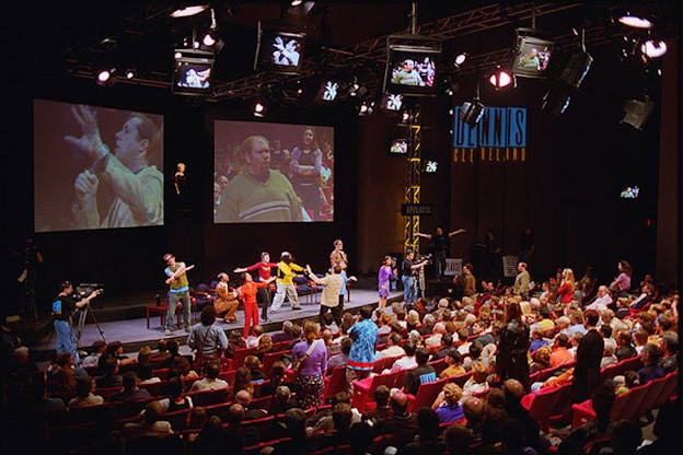 Several performers wearing clothes with primary colors stand on stage in front of an audience. On the sides of the stage, camera men point cameras into the audience. Two projectors show these members of the audience. One audience member speaks into a microphone and the other just stands and looks ahead. 