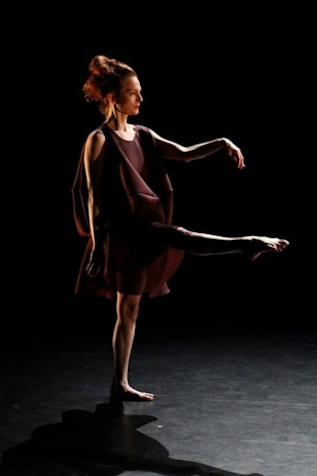 Performer on a dark stage wearing a flowy wine colored dress looks in the direction of their leg extended straight ahead and holds one bent arm parallel and the other resting at their side.
