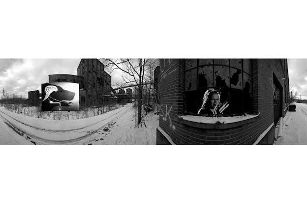 A black and white panorama image of buldings surrounded by snow. In the front right closest to the viewer an image of a figure wearing horns on top of their head and holding a cleaning supply in their hand is clogged on a building's broken window. 