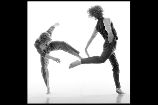 A black and white performance still of two dancers in a white space. The dancers wear all black and face away from the camera.  The dancer on the left leans forward over their right bent leg and lifts their left leg up by their waist. The dancer on the right bends backwards towards the left and balances on their left leg with their other leg and both arms behind them. 