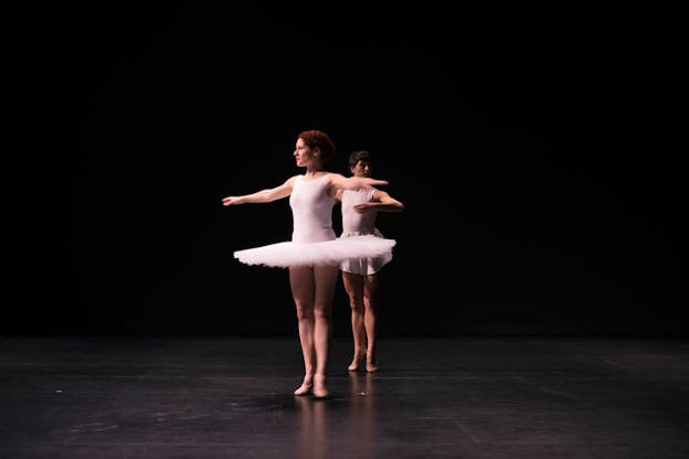 Two ballet dancers in white leotards and tutus relevé on a dimly lit stage.