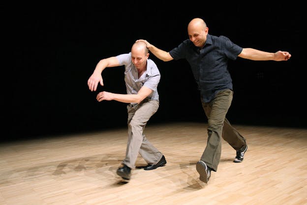 Two performers run, wearing khakis and short sleeve button down shirts. The person on the left lunges forward with their arms raised at chest level and slightly to their right. The person on the right smiles and has their right hand on the other person's head and their left arm extended out to their side. The performers walk on a wooden stage and the background is all black.