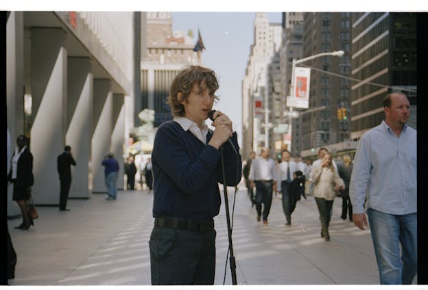 Sharon Hayes is centered in an image of a New York City sidewalk with pedestrians in the background. She stands and speaks into a microphone on a stand, dressed in a navy sweater over a white collared shirt, and dark green pants with a black belt. 