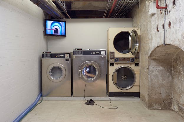 Four aligned laundromats. In the middle one a person has entered holding a microphone of which the cable hangs out and slithers behind the other machines. Next to the cable, two pair of slippers. 
