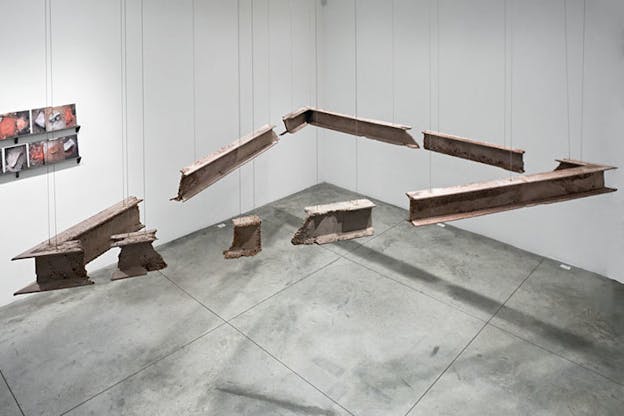 Fragments of rusty steel beams in desaturated red are hung slightly diagonally from the ceiling to form an obtuse triangle. The installation floats above a concrete floor against a white background. On the left of the image, there is a partial view of another artwork on the wall.
