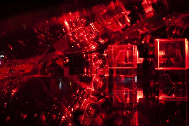 Spheres and cubes situated on a glistering dark surface illuminated by red light, the ceiling above them mirroring the shapes and reflecting the light.