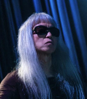 Keiji Haino looks up toward the right, bathed in blue light, his brow furrowed. He wears dark sunglasses and has long white-silver hair that falls over his shoulders.