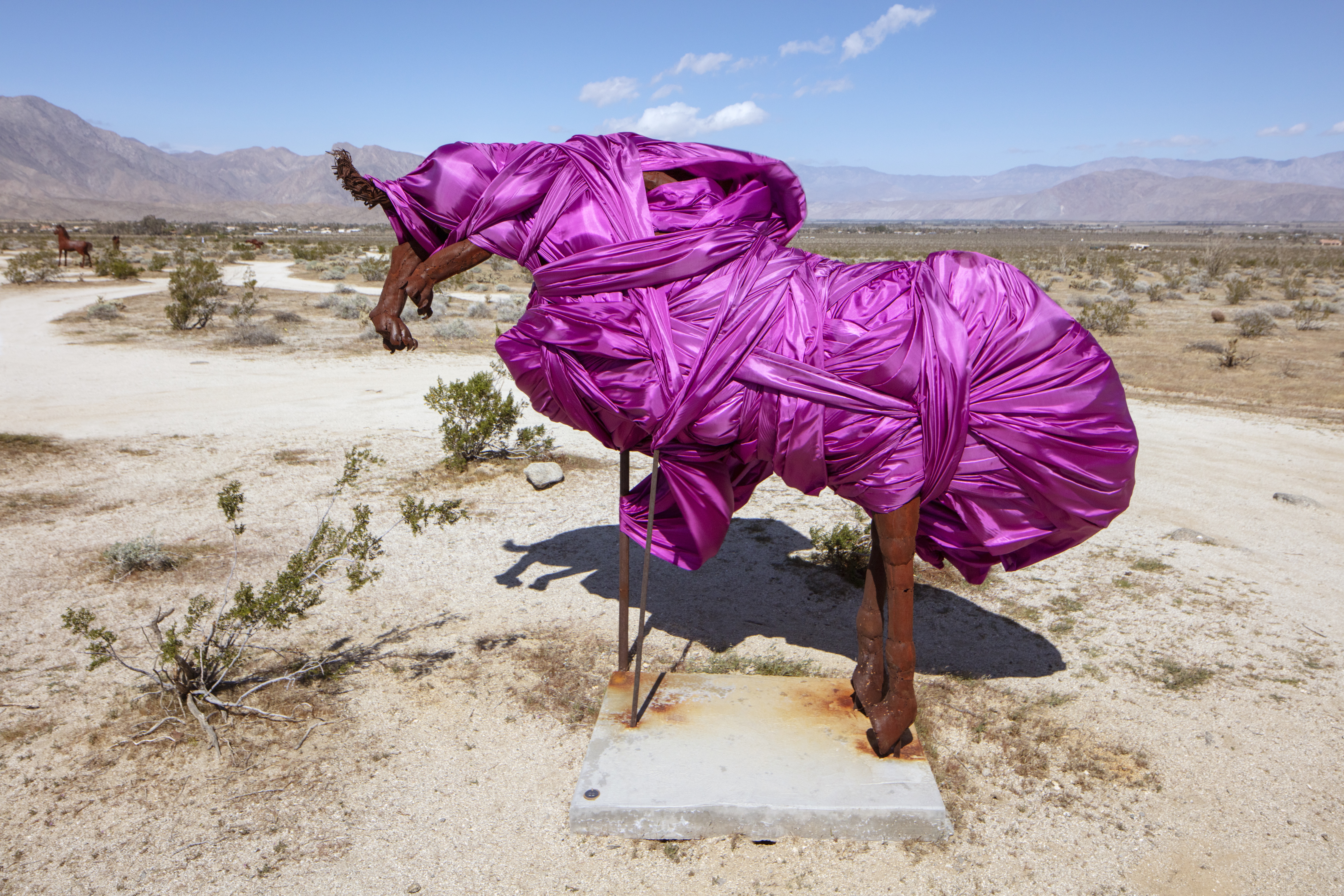 A red rust-colored sculpture that looks like the outline of a bucking horse being attacked by a predator with claws is wrapped in bright fuchsia fabric. In the background the desert extends into the horizon until it meets the mountains.