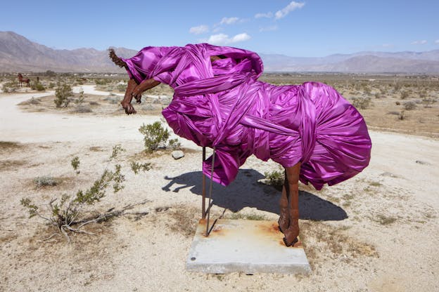 A red rust-colored sculpture that looks like the outline of a bucking horse being attacked by a predator with claws is wrapped in bright fuchsia fabric. In the background the desert extends into the horizon until it meets the mountains.