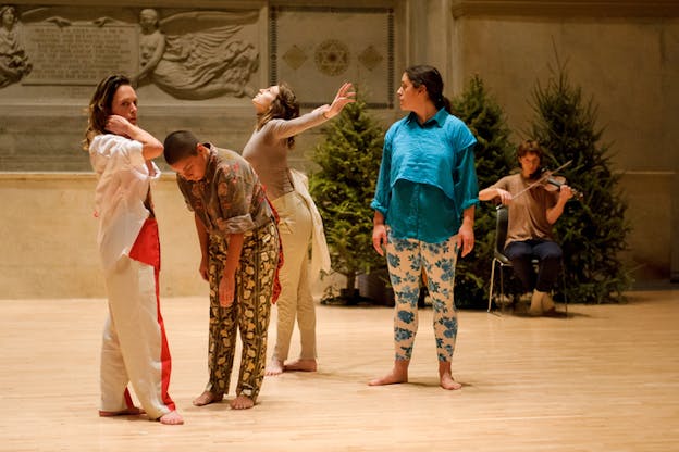 Four performers dressed in solid colors and floral prints stand on a wooden floor as a seated musician behind them plays the violin in front of pine trees.