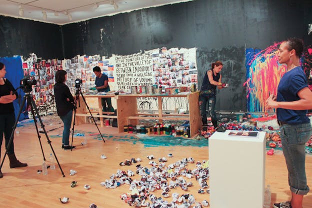 People appearing to be filmed in a room with paper balls on the floor, buckets and tubes of paint, and paint-splattered and text-pasted walls. 