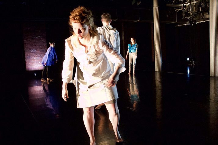 Performers stand on a black stage in various frozen stances. A performer in front dressed in a white dress with ginger hair closest to the viewer lifts one leg while their arms dangle to the sides.