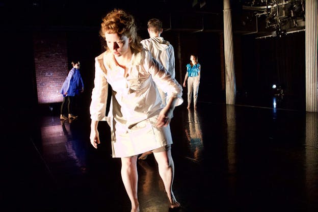 Performers stand on a black stage in various frozen stances. A performer in front dressed in a white dress with ginger hair closest to the viewer lifts one leg while their arms dangle to the sides.