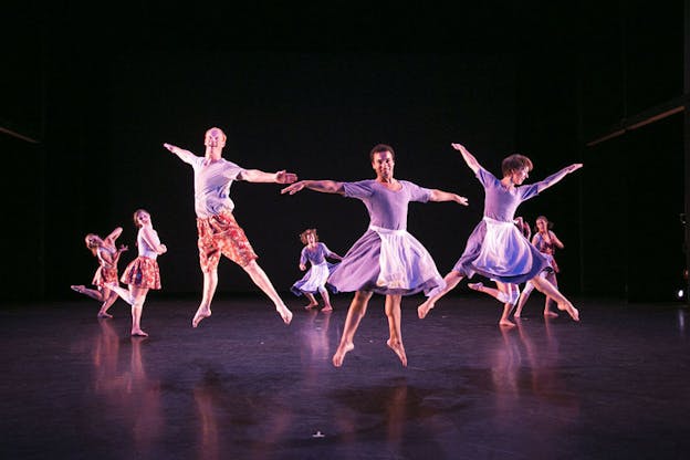 Performers in skirts in various pirouettes move in a circle. The three people in the front of the circle are smiling mid-air.