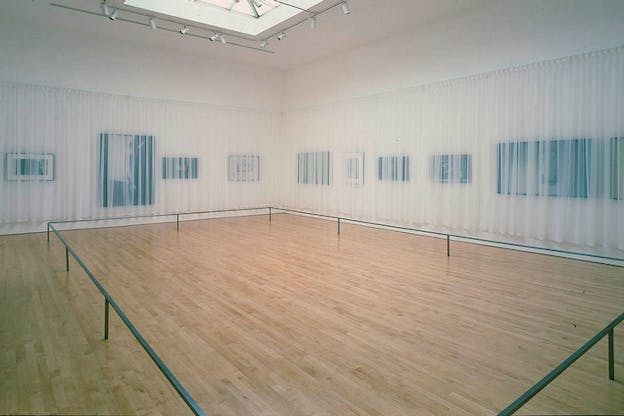 Polls close to the wooden floor form a rectangle. They are surrounding by dark canvases on the walls that are blurred behind transparent curtains. 