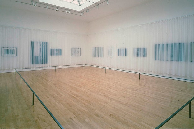 Polls close to the wooden floor form a rectangle. They are surrounding by dark canvases on the walls that are blurred behind transparent curtains. 