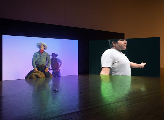 Two screens stand ca. The left screen shows two performers in cowboy garb, one riding a mechanical bull, and the right screen shows a performer wearing a white t-shirt in mid-movement.