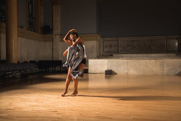 A performer dances by themselves in an empty space. A glimmering metallic silver scarf hangs from their neck forming different shapes by their movement. Behind them a marble structure stands on the wooden floorboard.