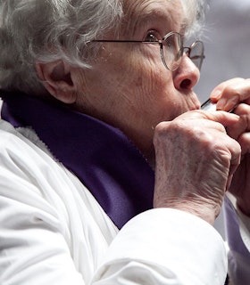 A close up portrait of Pauline Oliveros blowing into a harmonica. She has short grey hair and wears thin wire glasses, a purple scarf and a white shirt. 